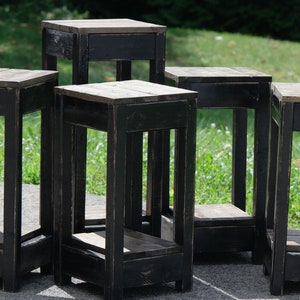 May include: Five black wooden end tables with a distressed finish. Each table has a shelf and a drawer. The tables are arranged in a circle on a green grassy lawn.