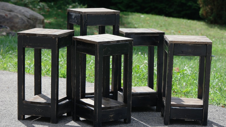 May include: Five black wooden end tables with a distressed finish. Each table has a shelf and a drawer. The tables are arranged in a circle on a green grassy lawn.