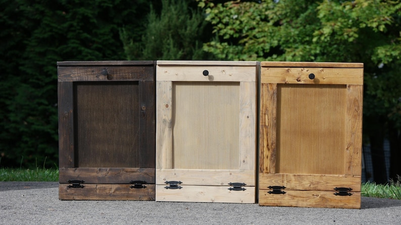 May include: Three wooden trash cans with hinged lids and black metal handles. The cans are stained in different shades of brown, from dark brown to light brown.
