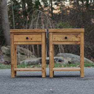 May include: Two matching wooden nightstands with a medium brown stain and visible wood grain. Each table features a single drawer with a small, round black knob and an open lower shelf supported by four straight legs with a rectangular top surface.