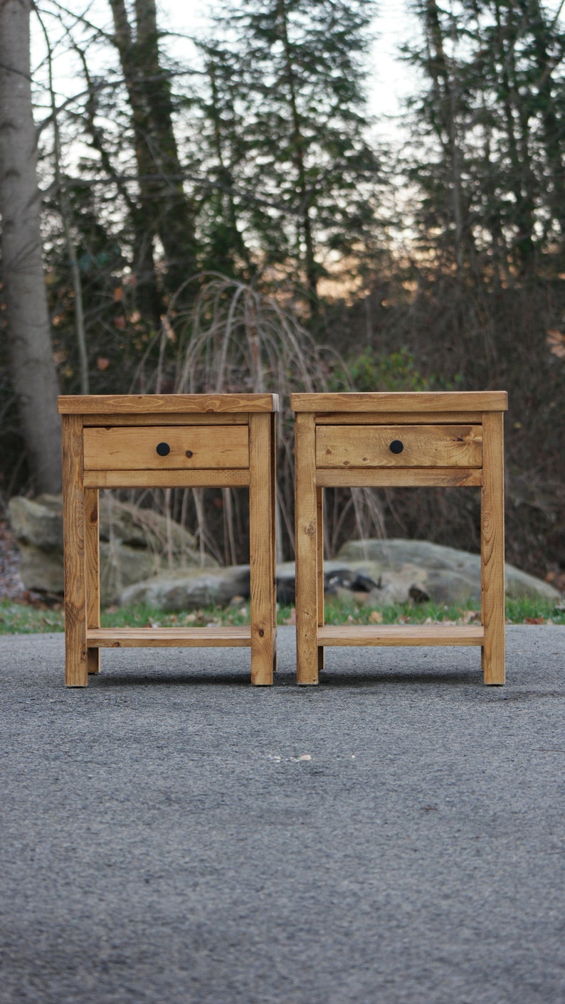 May include: Two matching wooden nightstands with a medium brown stain and visible wood grain. Each table features a single drawer with a small, round black knob and an open lower shelf supported by four straight legs with a rectangular top surface.