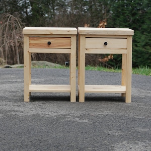 May include: Two light-colored wooden nightstands, each featuring a single drawer with a small, round black knob and an open lower shelf. The tables are constructed from natural wood with visible grain patterns and have a rectangular top surface.