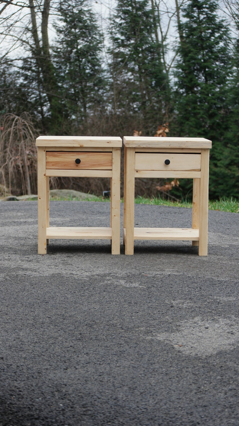 May include: Two light-colored wooden nightstands, each featuring a single drawer with a small, round black knob and an open lower shelf. The tables are constructed from natural wood with visible grain patterns and have a rectangular top surface.