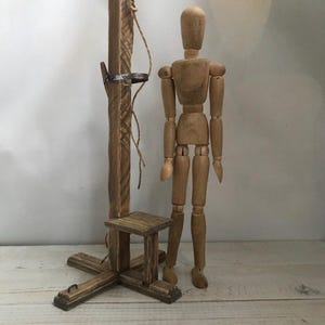 Könnte beinhalten: Eine Miniatur-Holzguillotine mit einem kleinen Sitz und einer hölzernen Künstlerpuppe. Die Guillotine ist aus gebeiztem Holz gefertigt und hat einen Metallklingenhalter. Die Puppe ist beweglich und aus hellem Holz gefertigt.