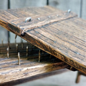 Puede incluir: Una tabla de madera con una tapa con bisagras, que revela un lecho de clavos debajo. Los clavos están dispuestos en filas y son de metal. La tabla es marrón y tiene un aspecto rústico.