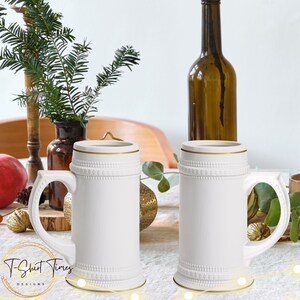 May include: Two white ceramic beer steins with gold trim, sitting on a white tablecloth with a string of lights. The steins have a raised design around the top and bottom.