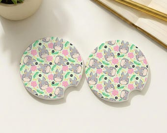 May include: Two round car coasters with a light green background, featuring a pattern of gray cartoon characters, pink flowers, and black accents. Each coaster has a curved notch for easy removal.