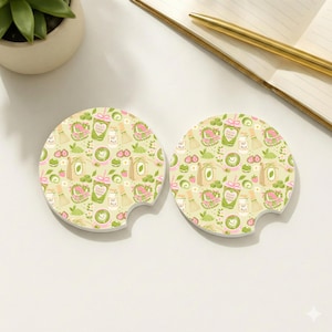 May include: Two round car coasters featuring a repeating pattern of matcha-themed illustrations, including green tea lattes, oat milk cartons, bamboo whisks, strawberries, and green tea cakes on a light cream background with a small notch on the side.