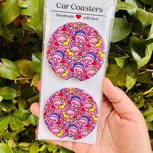 May include: Two circular car coasters in a clear plastic sleeve with a header card labeled Car Coasters Handmade with love. The coasters feature a repeating pattern of a pink cartoon character sleeping on a yellow star against a pink background.