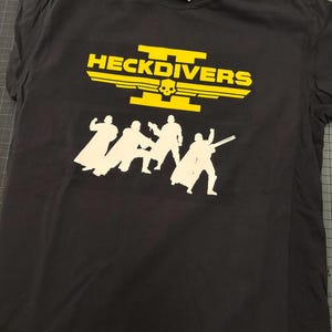 May include: Black t-shirt with a yellow graphic that reads "HECKDIVERS" above a skull and the silhouettes of five figures. The shirt has a crew neck and a tag at the neckline.
