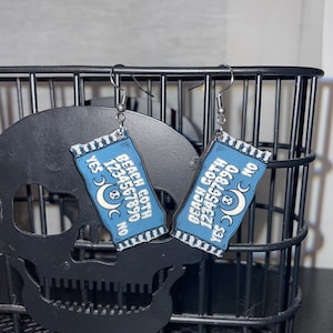 May include: A pair of blue and white dangle earrings with a beach goth theme. The earrings feature a crescent moon, a sun, and the numbers 1234567890. The text "Beach Goth" is printed on the earrings.