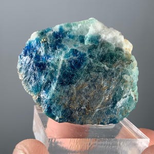 May include: A close-up of a vibrant blue and green mineral specimen. The stone has a rough, textured surface with areas of deep blue and lighter turquoise hues. It is displayed on a clear, rectangular stand against a gray background.