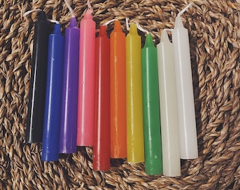May include: A collection of nine colorful taper candles arranged on a woven, circular surface. The candles are in various colors, including black, blue, purple, pink, red, orange, yellow, green, and white. Each candle has a white wick.