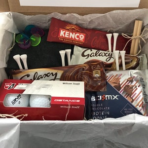 May include: A gift box filled with golf-themed items, including golf balls, tees, a chocolate bar, a coffee packet, and a protein cookie. The box is lined with tissue paper and has a handwritten note that says "just for you with love".