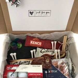 May include: A gift box filled with golf-themed items, including golf balls, tees, chocolate bars, and a protein cookie. The box is lined with brown paper and has a white card on top that says "just for you with love".