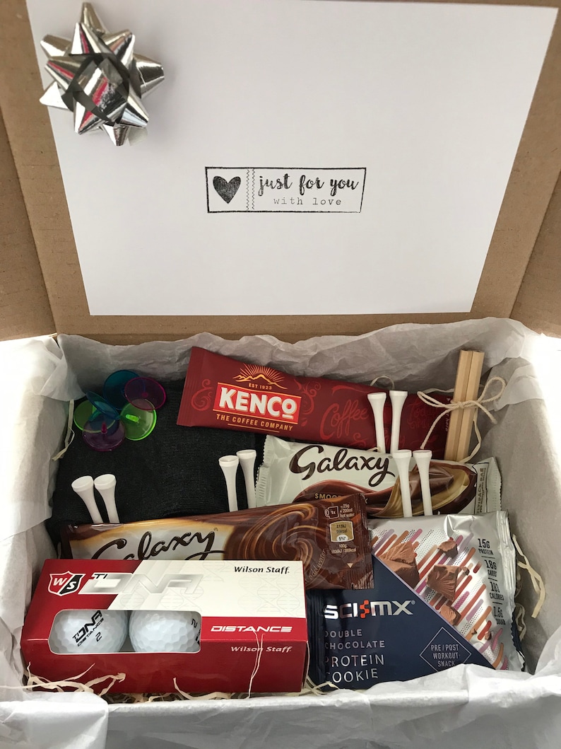 May include: A gift box filled with golf-themed items, including golf balls, tees, chocolate bars, and a protein cookie. The box is lined with brown paper and has a white card on top that says "just for you with love".
