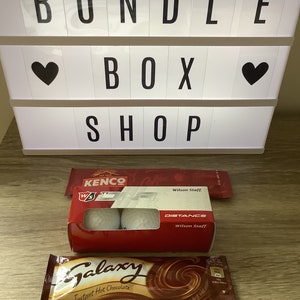 May include: A bundle box shop sign with a heart on each side. The sign reads "BUNDLE BOX SHOP". Below the sign is a box of golf balls with the text "KENCO" and "Wilson Staff" on the box. There is also a chocolate bar with the text "Galaxy Instant Hot Chocolate" on the wrapper.