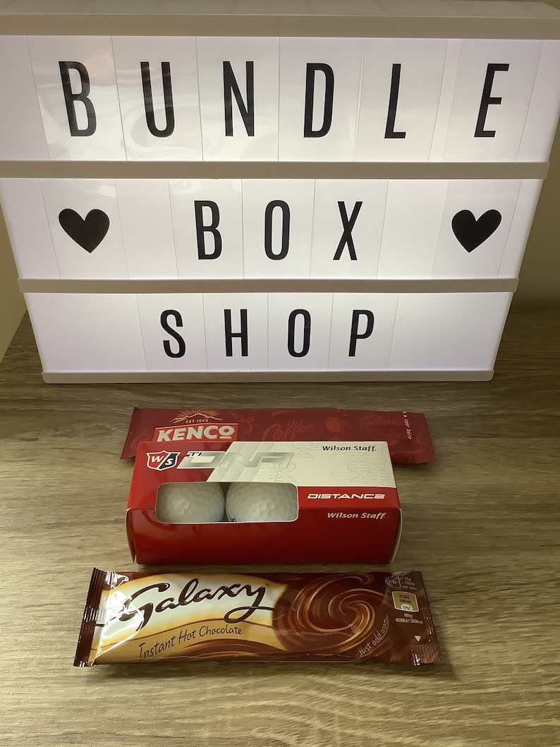 May include: A bundle box shop sign with a heart on each side. The sign reads "BUNDLE BOX SHOP". Below the sign is a box of golf balls with the text "KENCO" and "Wilson Staff" on the box. There is also a chocolate bar with the text "Galaxy Instant Hot Chocolate" on the wrapper.