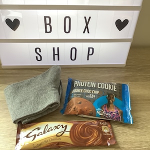 May include: A gray pair of socks, a brown chocolate bar with the text "Galaxy Instant Hot Chocolate", and a blue protein cookie package with the text "Phutein Cookie Double Choc Chip 23g Protein" and a picture of a person running.