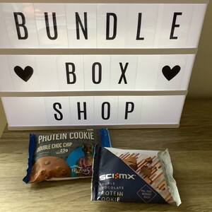 May include: Two protein cookie packages. One is blue and white with the text "Protein Cookie Double Choc Chip 23g Protein". The other is blue and white with the text "Sci-Mx Double Chocolate Protein Cookie Pre/Post Workout Snack".