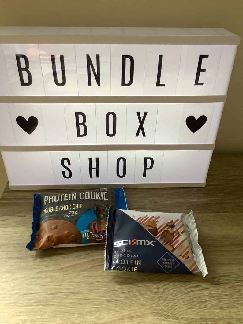 May include: Two protein cookie packages. One is blue and white with the text "Protein Cookie Double Choc Chip 23g Protein". The other is blue and white with the text "Sci-Mx Double Chocolate Protein Cookie Pre/Post Workout Snack".