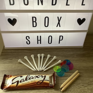 May include: A bundle box shop sign with the words "Bundle Box Shop" in black letters on a white background. The sign also has two black hearts on either side of the word "Box". Below the sign are various items including a chocolate bar with the words "Galaxy Smooth Milk" on the wrapper, a set of white golf tees, and a set of colorful plastic discs.