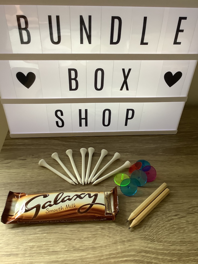 May include: A bundle box shop sign with the words "Bundle Box Shop" in black letters on a white background. The sign also has two black hearts on either side of the word "Box". Below the sign are various items including a chocolate bar with the words "Galaxy Smooth Milk" on the wrapper, a set of white golf tees, and a set of colorful plastic discs.
