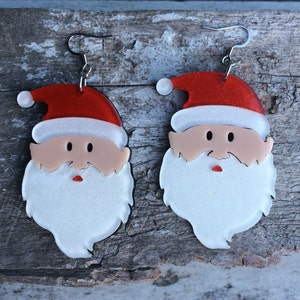 May include: A pair of Santa Claus-shaped earrings. Each earring features a red hat with a white pom-pom, a light skin-toned face, black eyes, a red mouth, and a white beard. The earrings are made of a shiny material and have silver-coloured hooks.