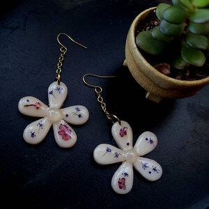 May include: A pair of white flower-shaped earrings with gold chains and hooks. The earrings are decorated with pressed flowers in shades of purple, pink, and white.