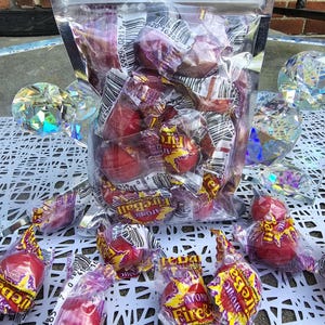 Pode incluir: Um saco de plástico transparente cheio de doces de canela Fireball vermelhos e roxos. Os doces são embalados individualmente em plástico transparente com etiquetas amarelas e pretas que dizem "Atomic Fireball".