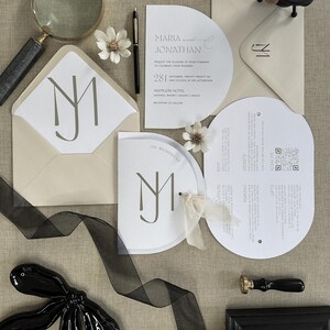 May include: A stylish wedding invitation suite with a semi-circular design, featuring the names "Maria and Jonathan". The set includes envelopes, RSVP cards, and a wax seal. The colour scheme is neutral with black accents and floral decorations.