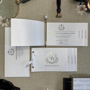 May include: A white wedding invitation with gold foil detailing. The invitation features a crest with the initials "DH" and the date "02.05.26". The text reads "Danielle & Harrold".
