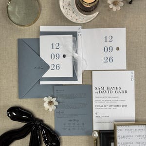 May include: A wedding invitation suite with a grey envelope, white cardstock, and a blue ribbon. The invitation features the date "Friday 12th September 2026" and the names "Sam Hayes and David Carr".