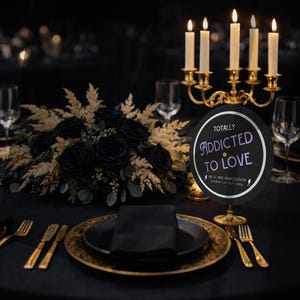 Op de afbeelding: Een romantische tafelsetting met zwarte rozen, gouden accenten en een bordje met de tekst "TOTALLY ADDICTED TO LOVE". Een kandelaar met brandende kaarsen en elegant glaswerk maken de scène compleet. De sfeer is romantisch en verfijnd.