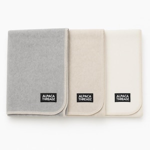 Op de afbeelding: Drie opgevouwen alpaca wollen dekens in grijstinten, beige en wit. Elke deken heeft een gestikte rand en een zwart label met de tekst "ALPACA THREADZ". De dekens zijn netjes opgestapeld.