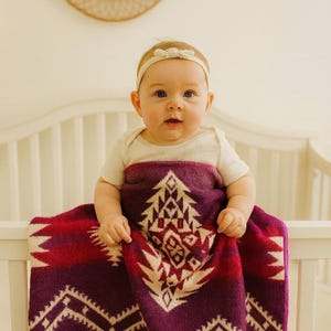 Può includere: Un bambino avvolto in una coperta a fantasia viola, rossa e crema. La coperta presenta un design geometrico con un motivo ad albero. Il bambino indossa un body bianco e una fascia bianca con un fiocco.