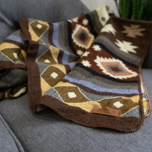 May include: A brown, yellow, and gray patterned throw blanket with a leather tag that says "Alpaca Apparel".