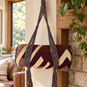 May include: A dark gray macrame plant hanger or strap featuring intricate knotted patterns and long tassels, suspending a rolled-up blanket with a geometric pattern in shades of deep burgundy, cream, and brown, hanging from a wooden wall hook.