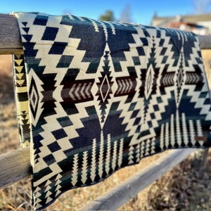 May include: A folded blanket with a geometric pattern in shades of cream, navy blue, black, and brown. The blanket is draped over a wooden fence, showcasing its intricate design. The pattern includes diamond and zigzag shapes.