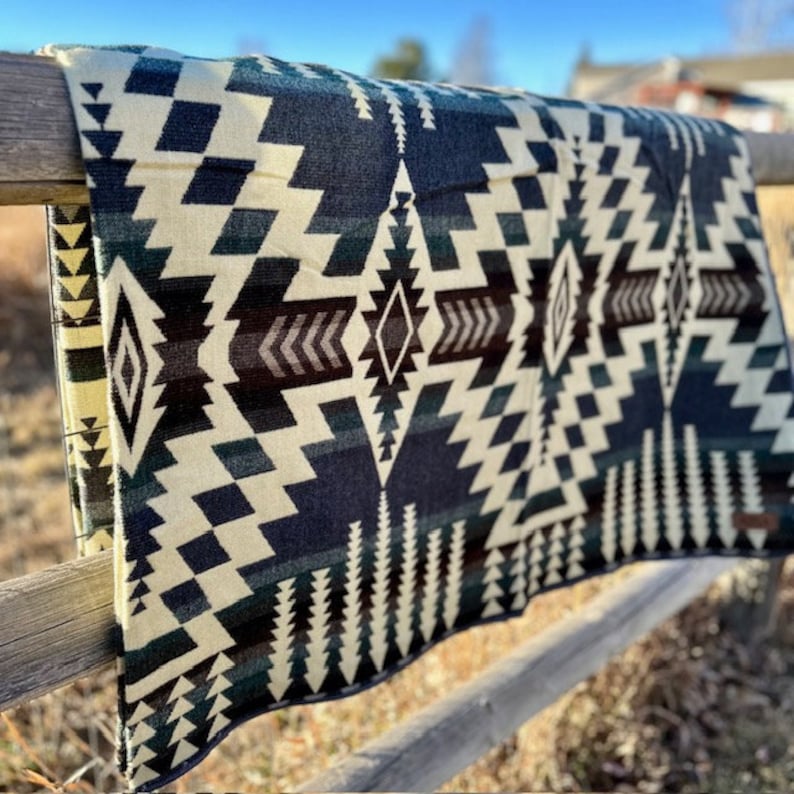 May include: A folded blanket with a geometric pattern in shades of cream, navy blue, black, and brown. The blanket is draped over a wooden fence, showcasing its intricate design. The pattern includes diamond and zigzag shapes.