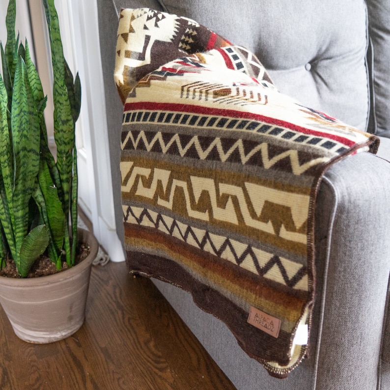 May include: A woven blanket with a geometric pattern in shades of brown, cream, and red is draped over a gray armchair. The blanket features a variety of shapes and lines, with a label that reads "Alpaca Threadz". A potted snake plant is in the background.