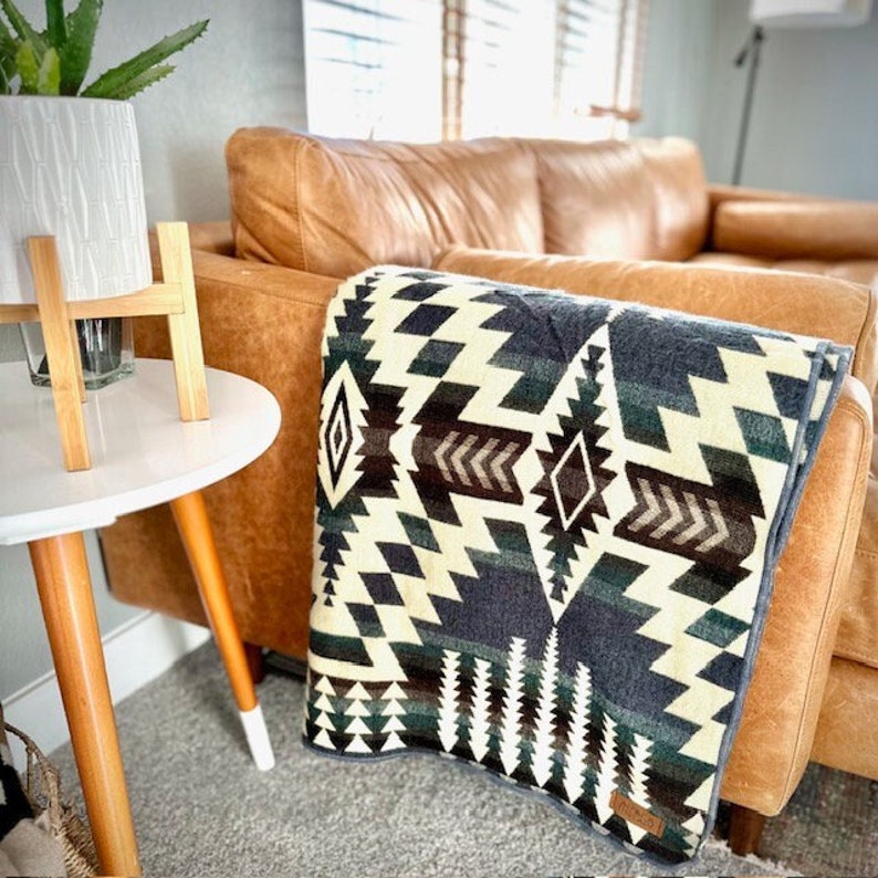 May include: A folded blanket with a geometric pattern in shades of cream, navy, green, and brown rests on a brown leather sofa. The blanket features diamond and zigzag designs. A white side table with wooden legs and a potted plant are in the foreground.