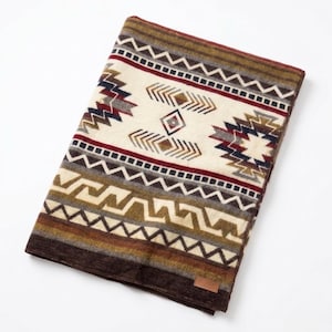 May include: A folded blanket with a Southwestern geometric pattern. The blanket features a cream-colored central panel with brown, red, and navy blue accents. The border is a dark brown, and the blanket has a small brown tag.