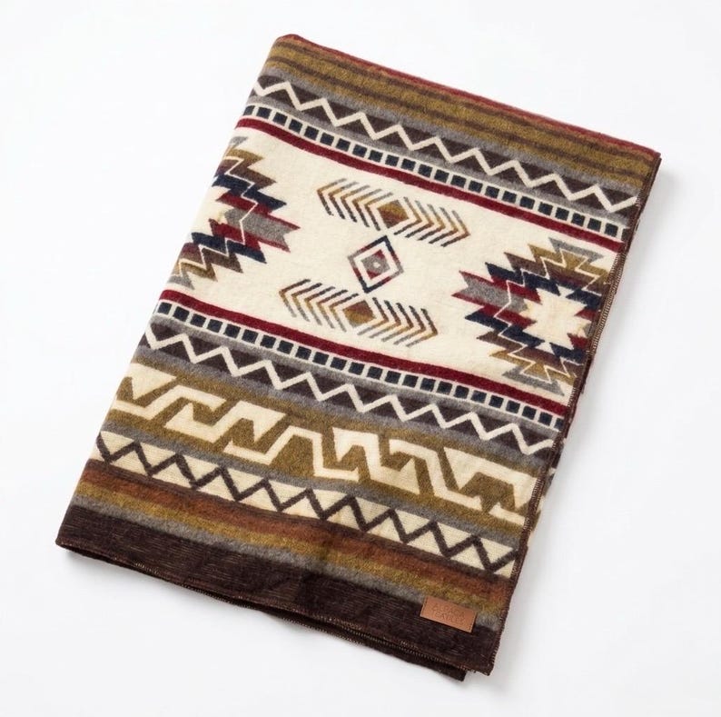May include: A folded blanket with a Southwestern geometric pattern. The blanket features a cream-colored central panel with brown, red, and navy blue accents. The border is a dark brown, and the blanket has a small brown tag.