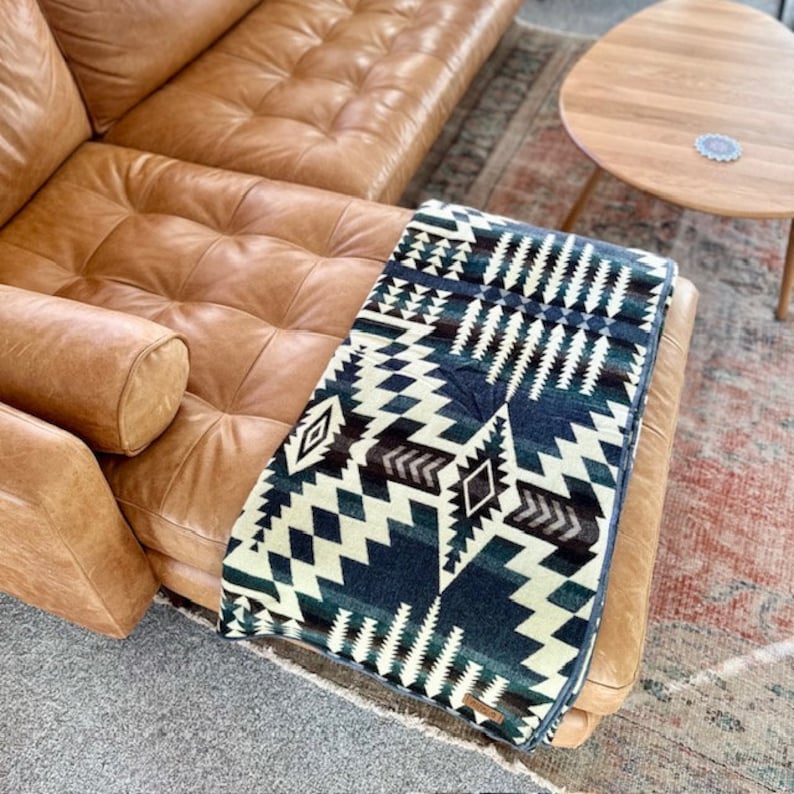 May include: A folded blanket with a geometric pattern in shades of blue, cream, and brown rests on a brown leather couch. The blanket features diamond and zigzag designs. The couch has a tufted design and is in a living room setting.