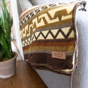 May include: A folded blanket with a geometric pattern in shades of brown, cream, and gray, draped over a gray armchair. The blanket has a leather tag that reads "ALPACA THREADZ".