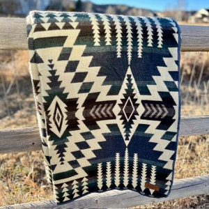 May include: A folded blanket with a geometric pattern in shades of cream, navy blue, and dark green. The design features diamond and zigzag shapes. The blanket is draped over a wooden fence in an outdoor setting.