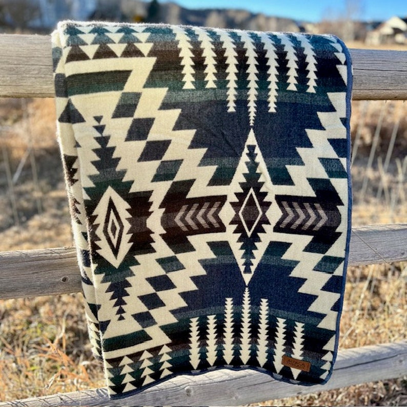May include: A folded blanket with a geometric pattern in shades of cream, navy blue, and dark green. The design features diamond and zigzag shapes. The blanket is draped over a wooden fence in an outdoor setting.