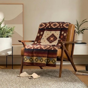 May include: A brown and tan patterned throw blanket draped over a mid-century modern chair with a leather seat. The blanket features geometric designs in shades of red, blue, and cream. Beige slippers rest on a gray rug.