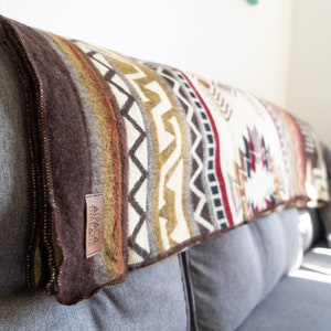 May include: A folded, patterned blanket with a brown border and a label that reads "ALPACA THREADZ." The blanket features a geometric design in shades of brown, cream, orange, and red. The blanket is draped over a gray sofa.